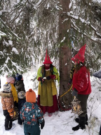 Metsätontut ovat lapsiryhmän kanssa kuusen juurella tutustumassa seuraavaan seikkailun tehtävään. Toinen tonttu lukee ohjeita ryhmälle.